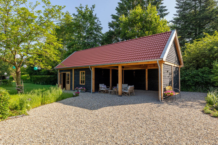 zadeldak-xxl-met-rode-dakpannen-zwarte-houten-wanden-en-overkapping-met-zitgedeelte-in-een-landelijke-tuin