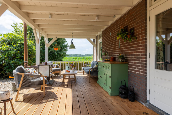 ruime-houten-veranda-aan-een-bakstenen-woning-met-houten-terrasvloer-zitmeubels-en-uitzicht-op-een-groene-tuin