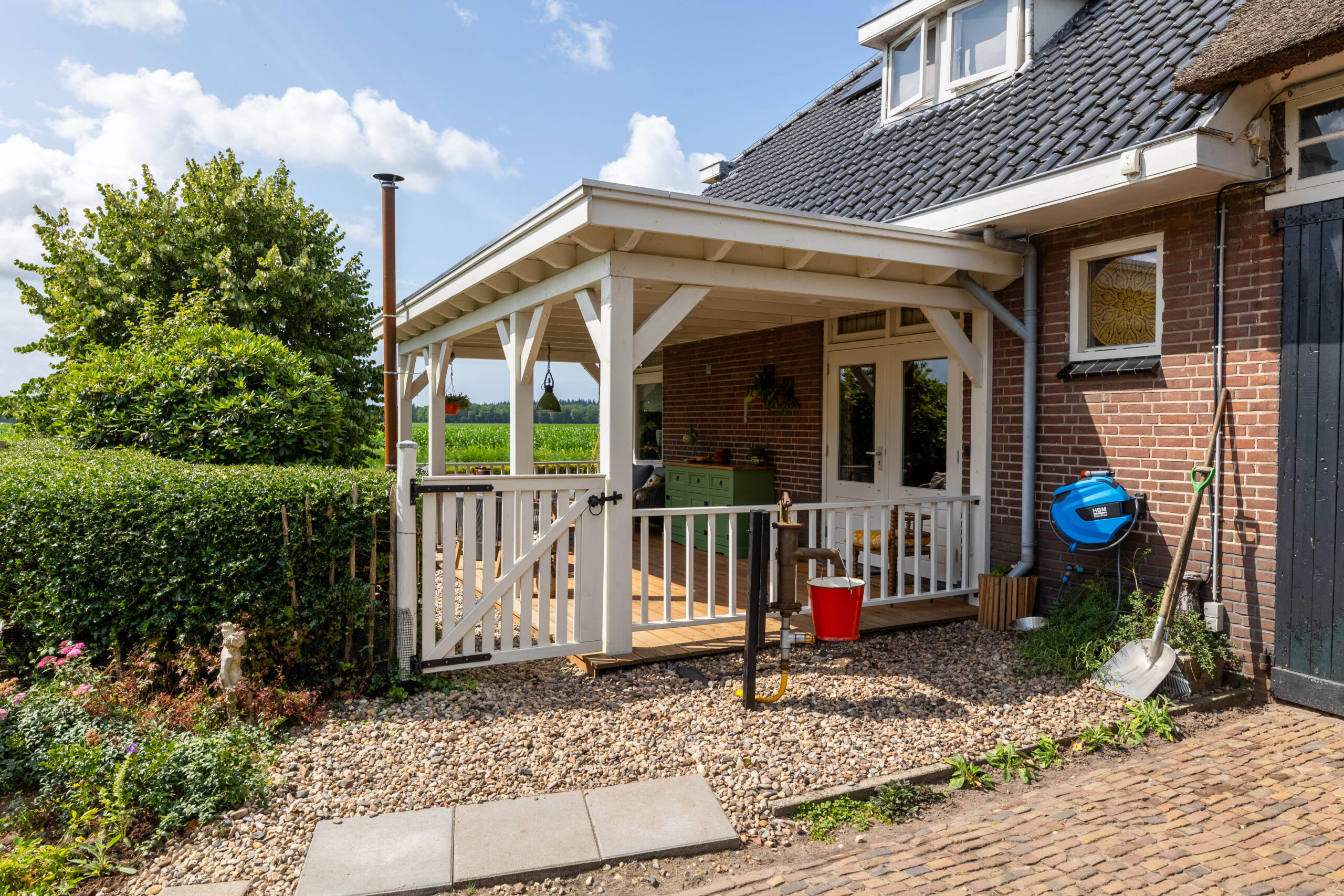 Landelijke-witte veranda-aan-een-bakstenen-woning-met-houten-hekwerk-en-houten-vlondervloer-in-een-groene-tuinomgeving.
