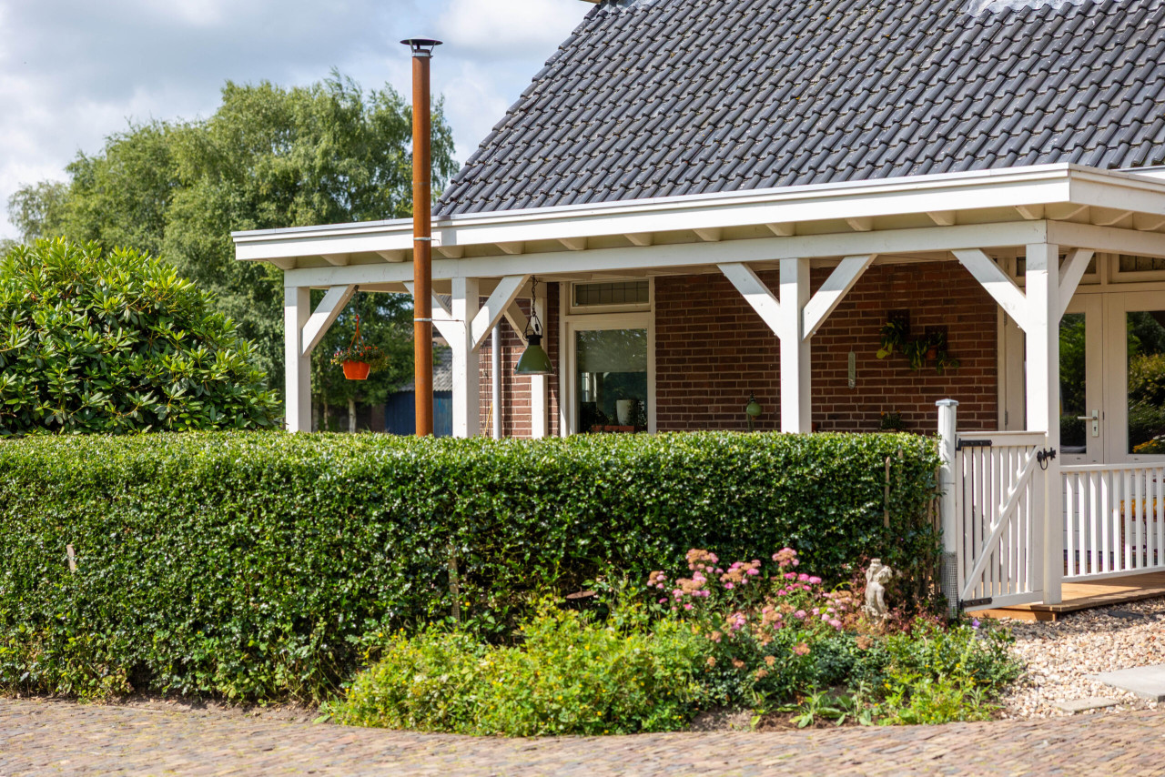 landelijke-witte-veranda-aan-een-bakstenen-woning-met-houten-constructie-en-hekwerk-gelegen-in-een-groene-tuin-met-haag