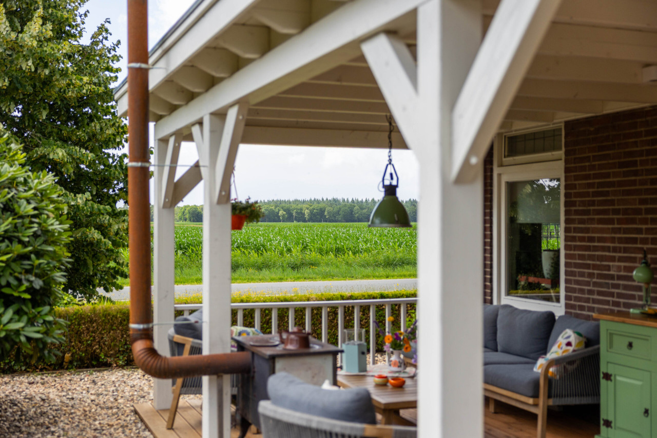 landelijke-witte-veranda-met-houten-constructie-en-zitgedeelte-geplaatst-aan-een-bakstenen-woning-met-uitzicht-over-een-groen-landschap