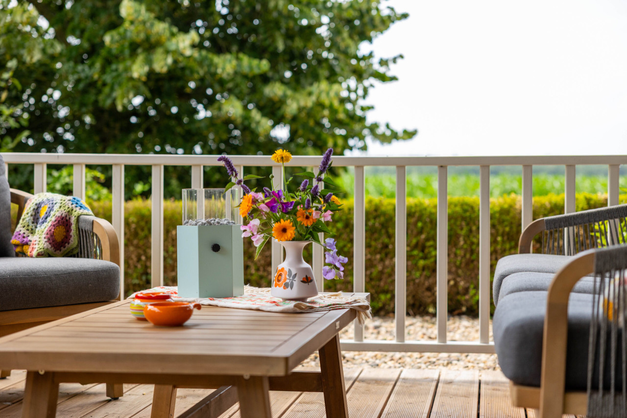 sfeervol-ingerichte-veranda-met-houten-terrasvloer-lounge-stoelen-salontafel-en-bloemenvaas-met-uitzicht-op-een-groene-tuin