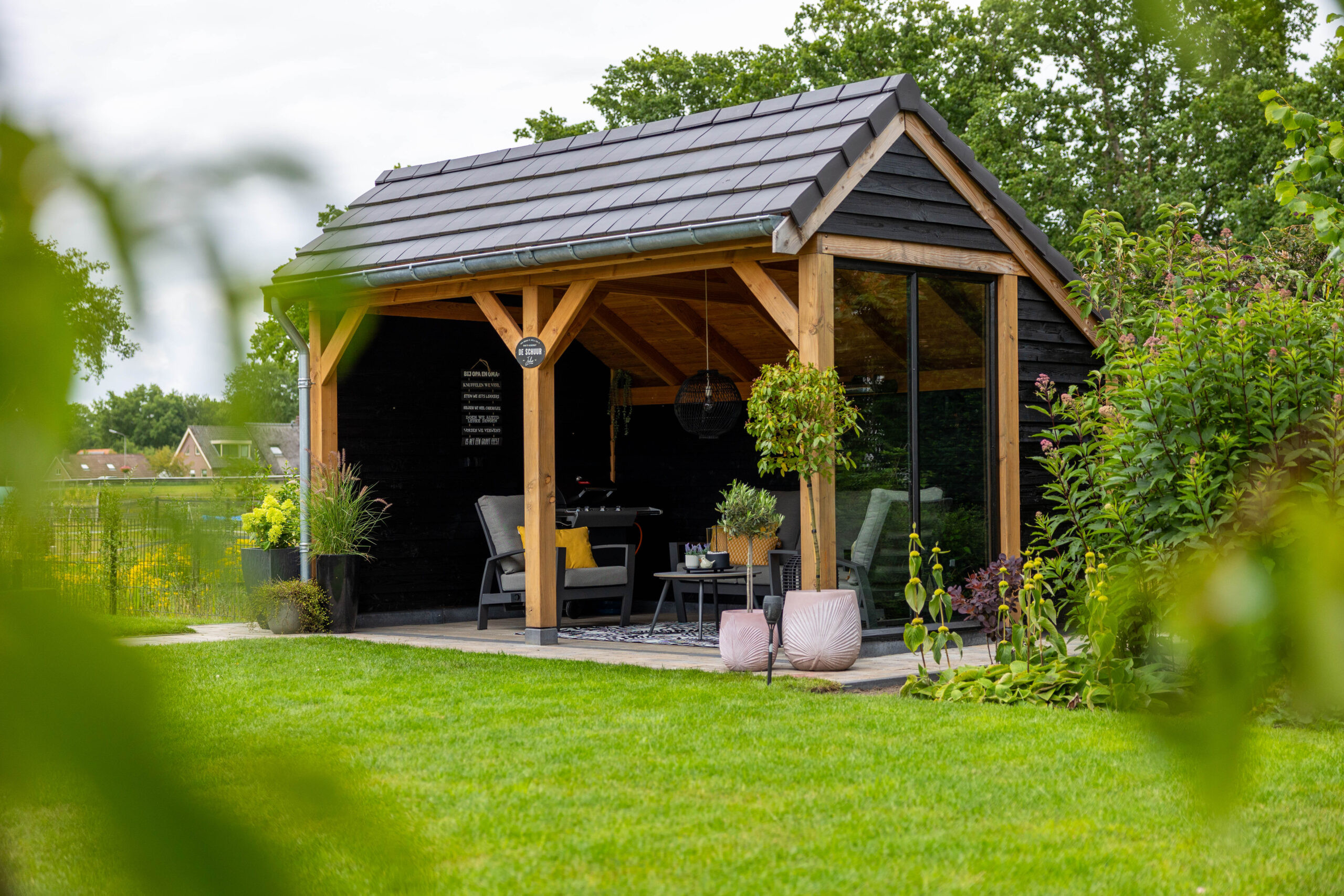 landelijke-houten-kapschuur-met-open-overkapping-in-een-groene-tuin-met-douglas-houten-constructie-zwarte-potdeksel-planken-en-donkere-dakpannen