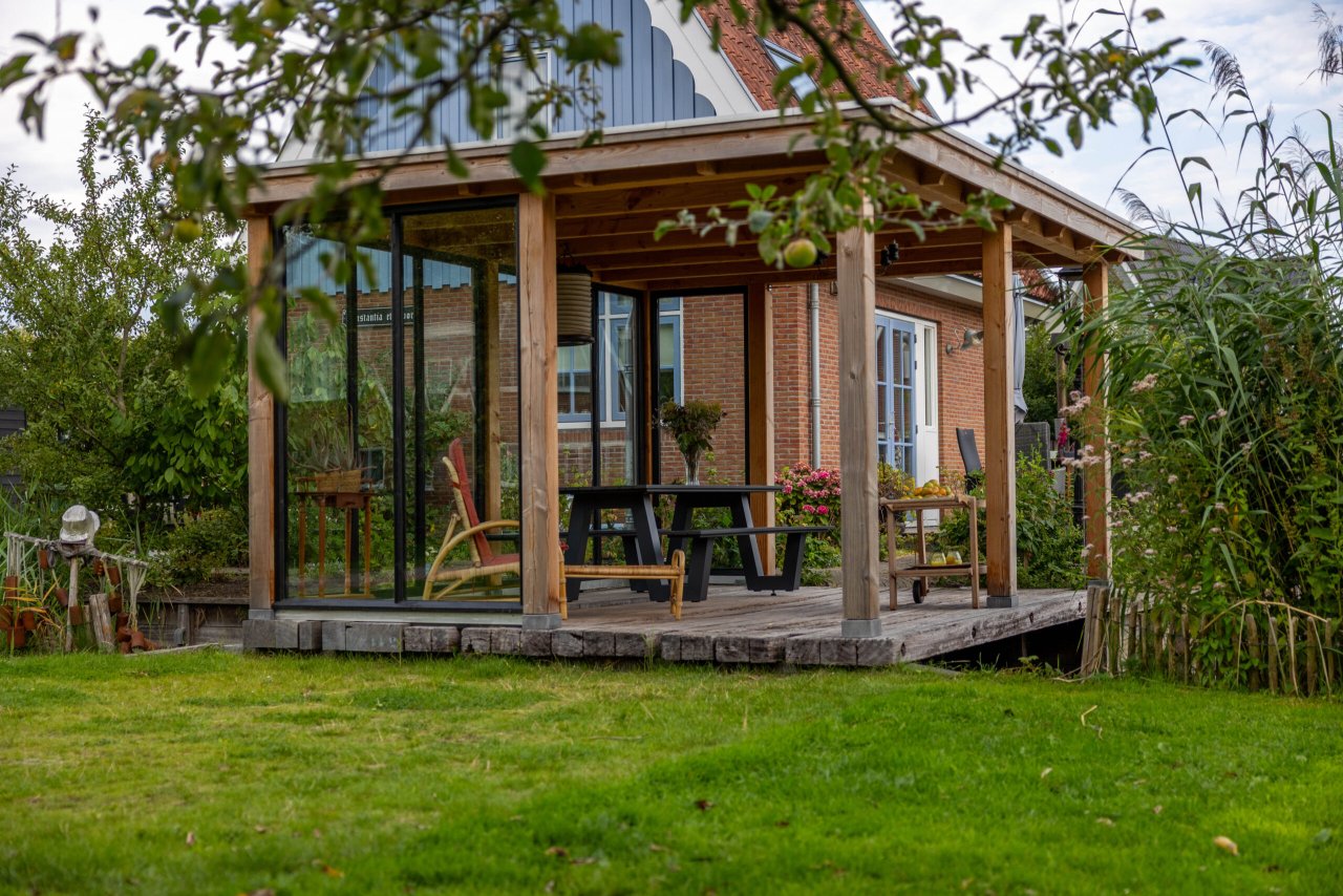 tuinkamer-overkapping-siena-op-houten-vlonder-met-glazen-wanden-in-groene-tuin-bij-het-water