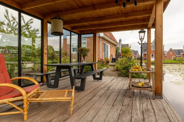 binnenkant-van-tuinkamer-overkapping-siena-met-houten-vlonder-tafel-met-banken-glazen-wanden-en-plantenrek-met-bloemen