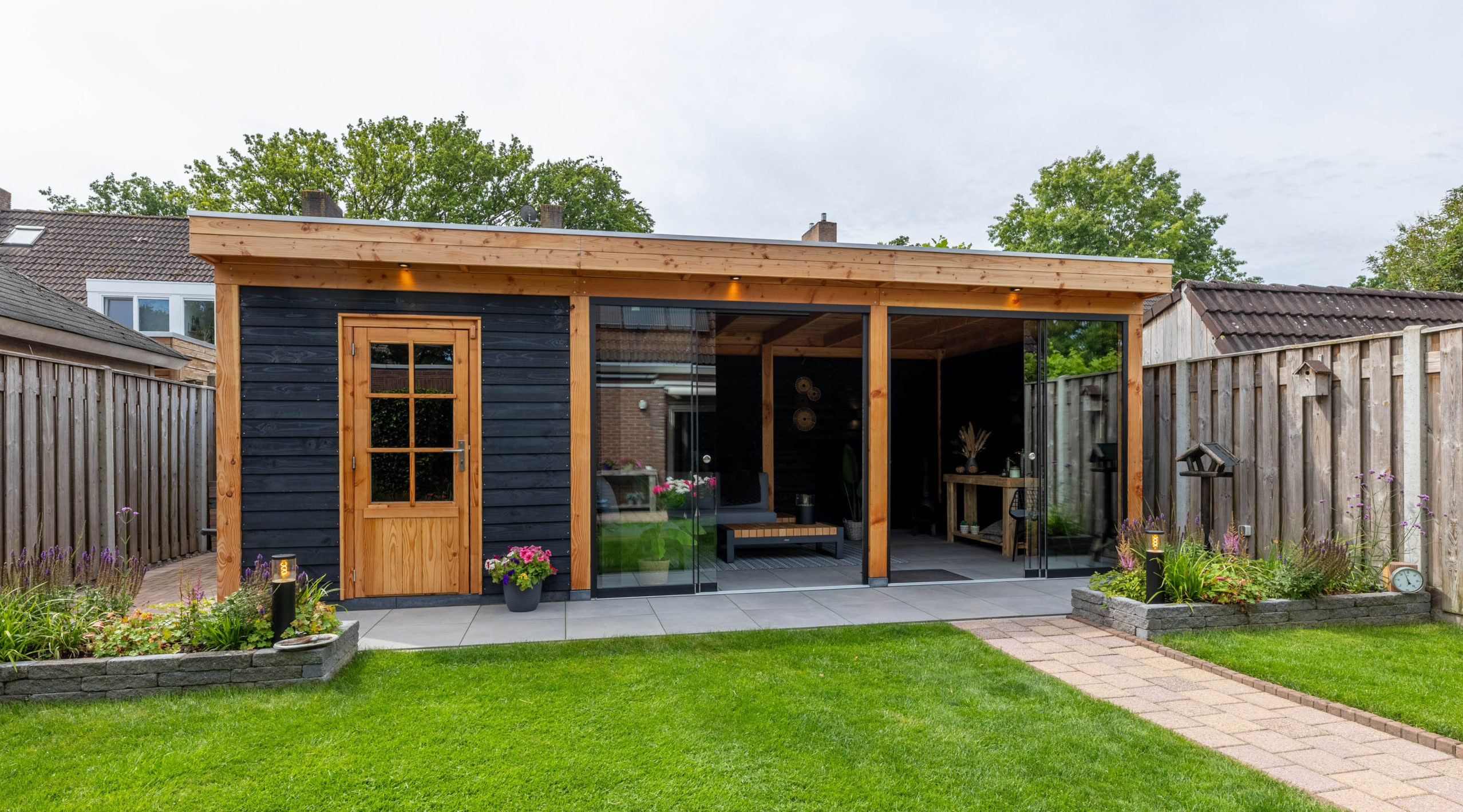 modern-houten-tuinhuis-met-opbergruimte-grote-glazen-schuifpui-en-overkapping-in-een-strakke-tuin